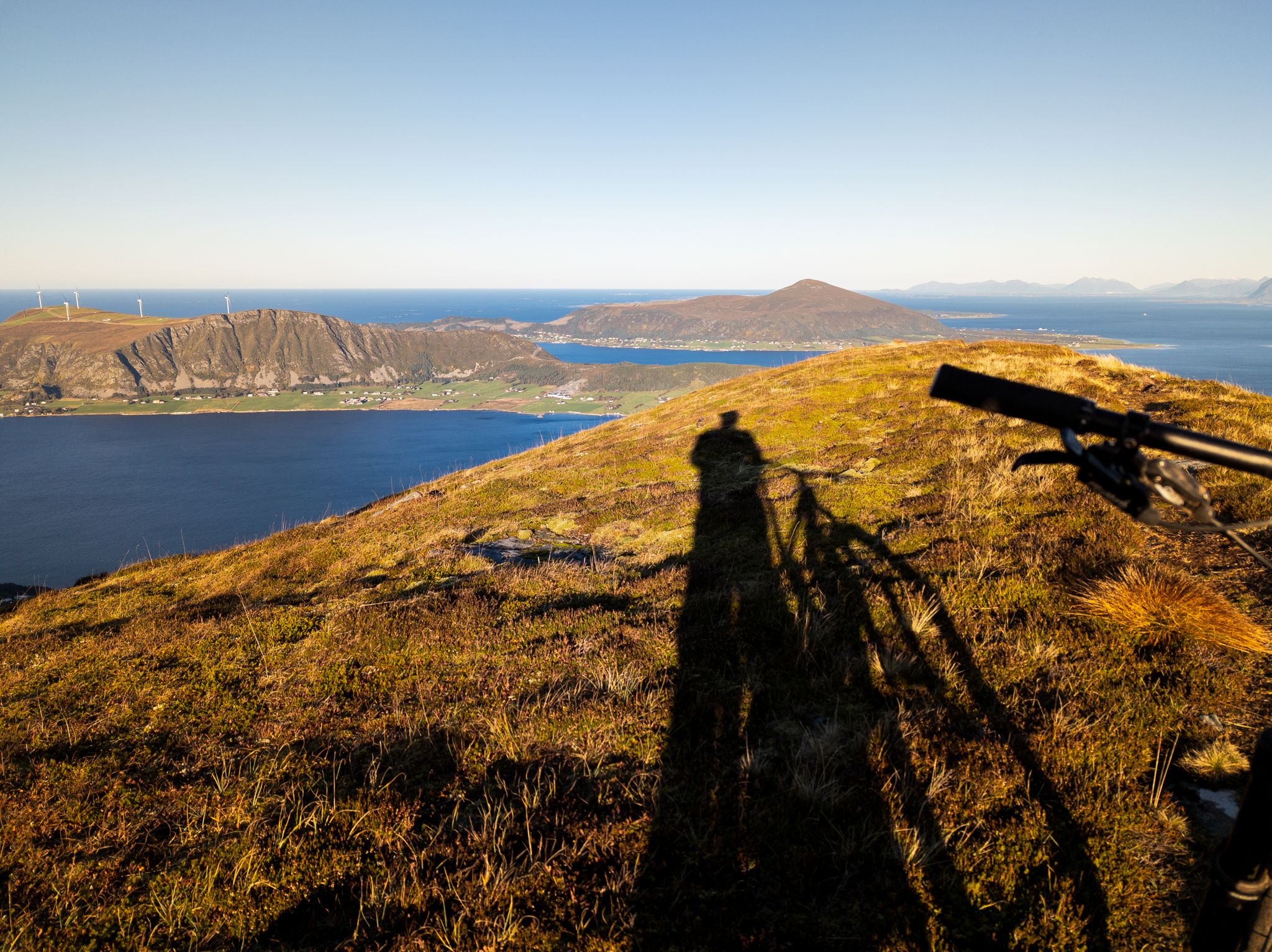 Sykkelstyre og lang skygge i lyngen med hav, øyer og vindturbiner i bakgrunnen på Lepsøya
