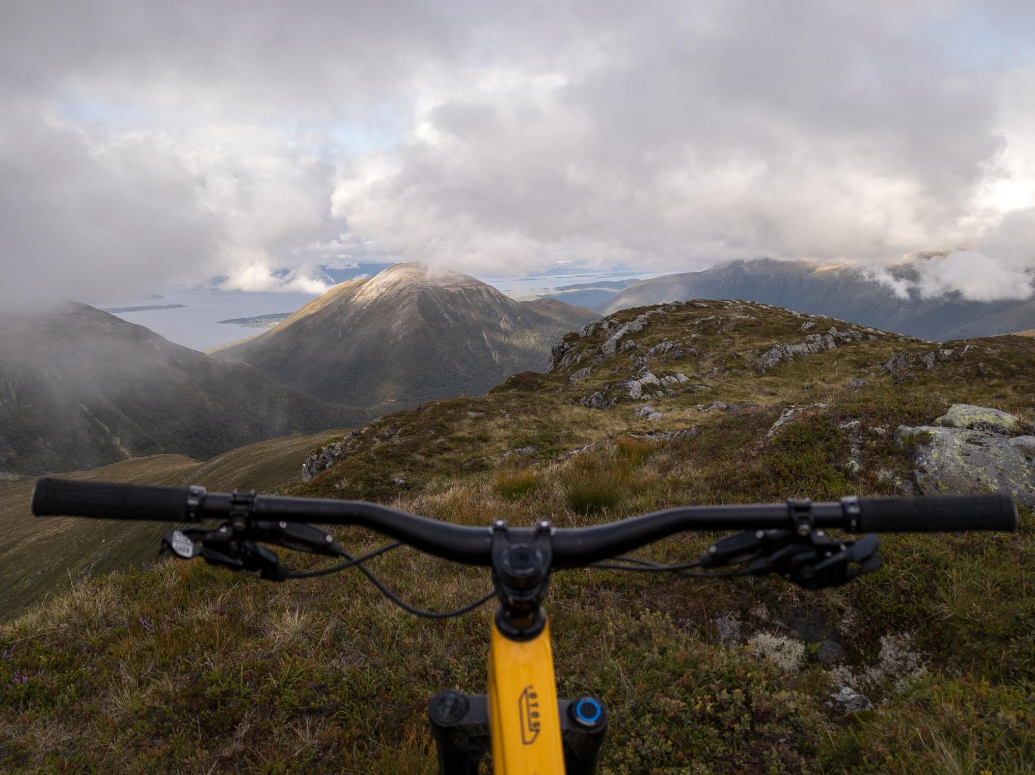 Gult sykkelstyre i forgrunnen over åpent fjellterreng, med skyer, fjord og fjell i bakgrunnen