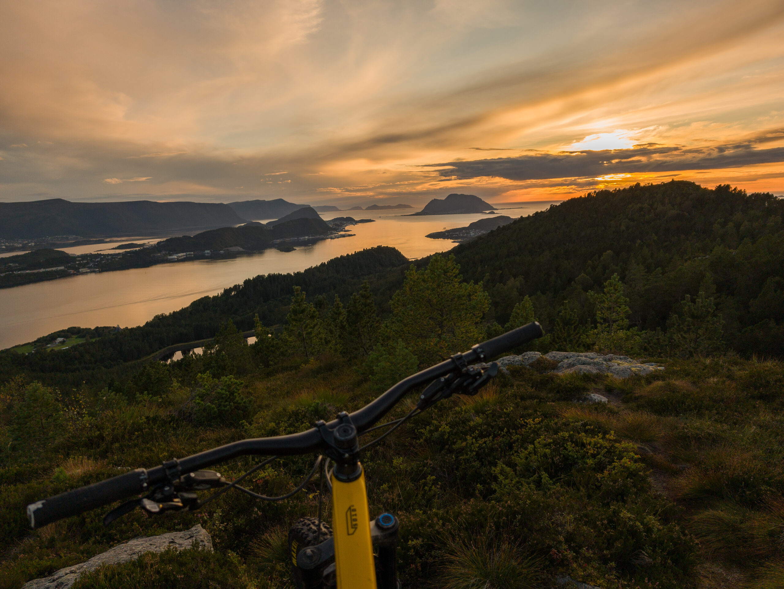 Styret på en gul terrengsykkel foran utsikten over fjord, øyer og kveldssol fra Myklebusthornet.