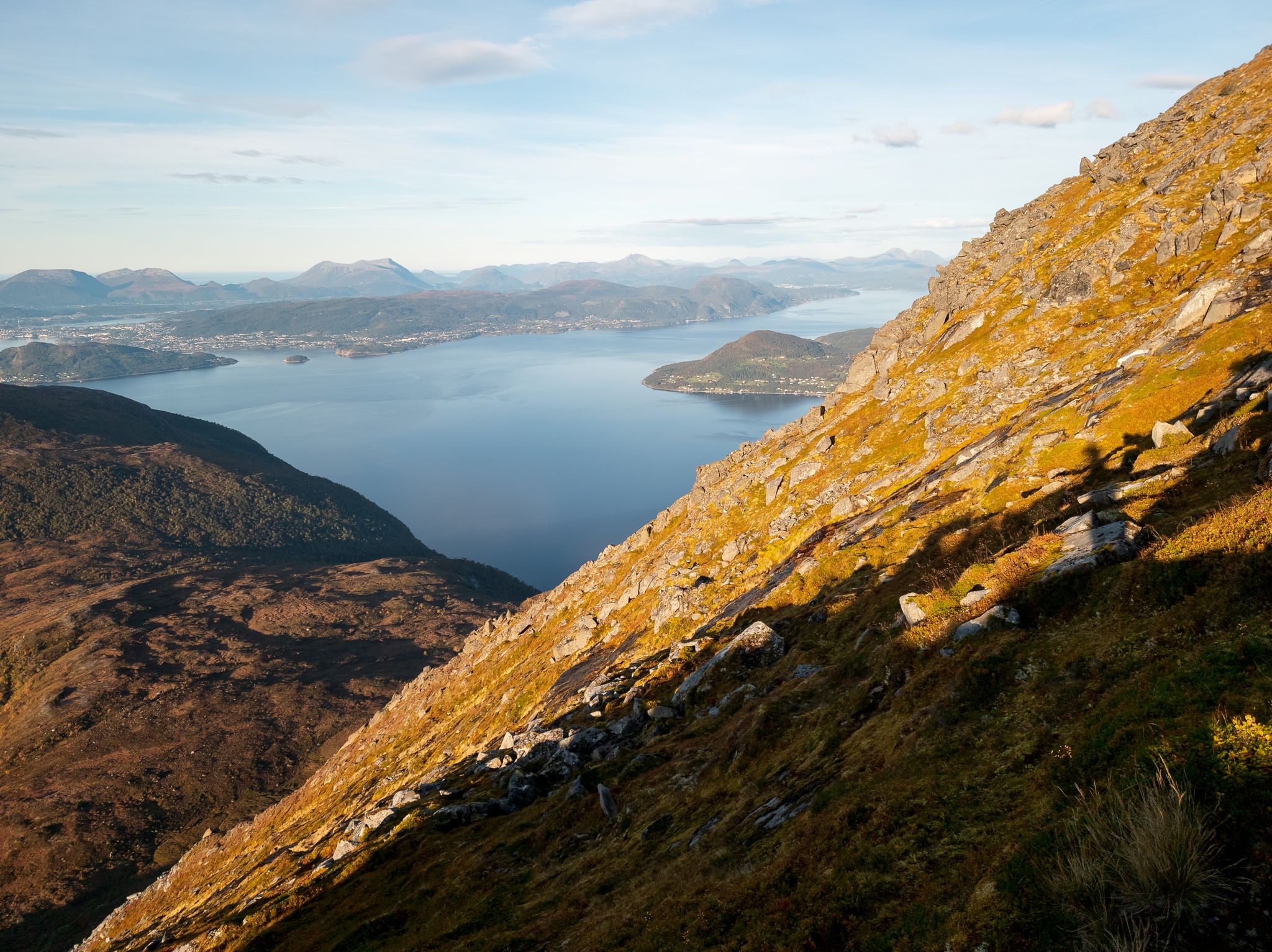 Bratt gress- og steinside i kveldssol med fjord og fjell i bakgrunnen