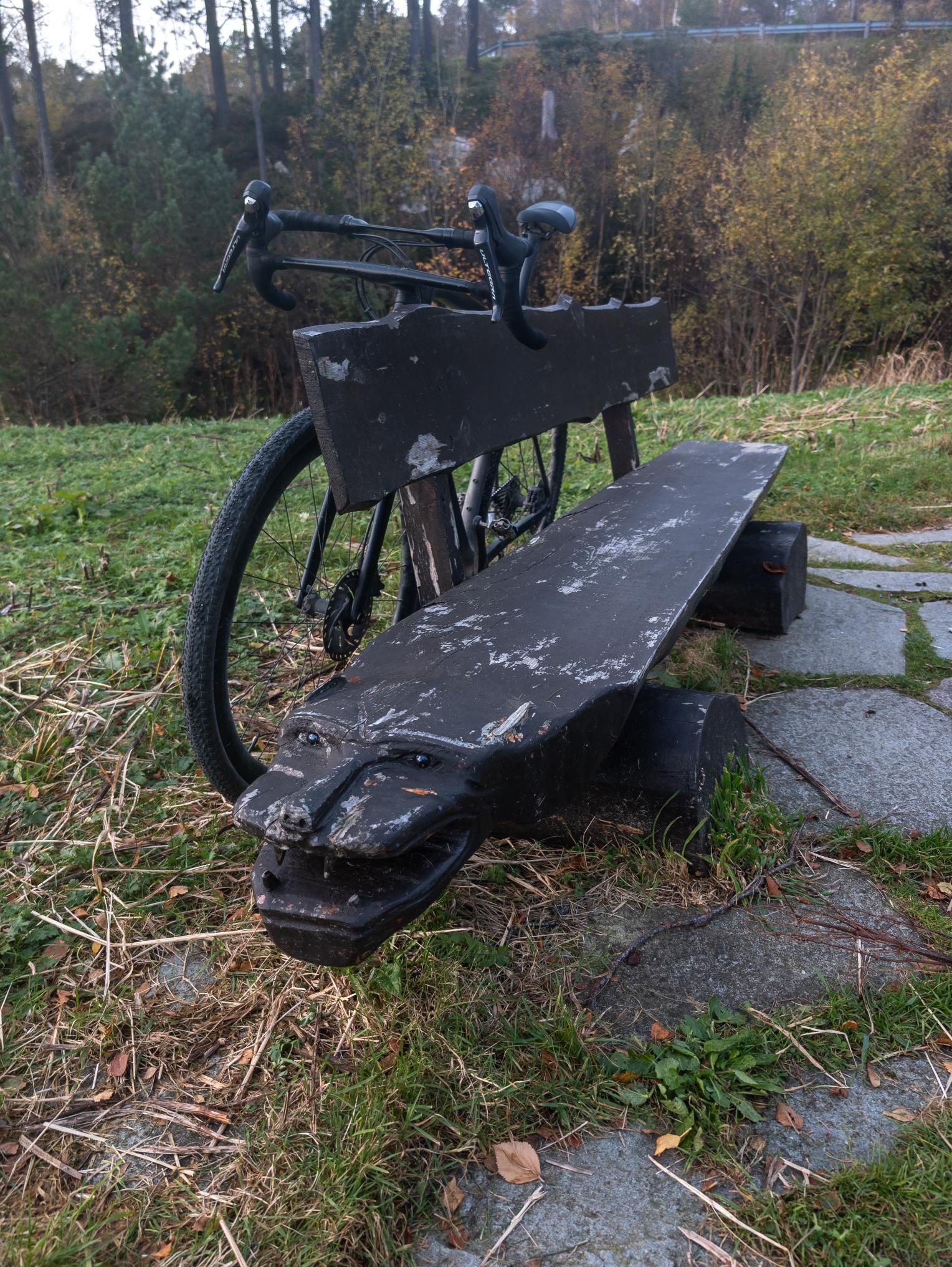 Dyreformet trebenk ved en steinlagt sti, med en sykkel bak benken og høstskog i bakgrunnen.