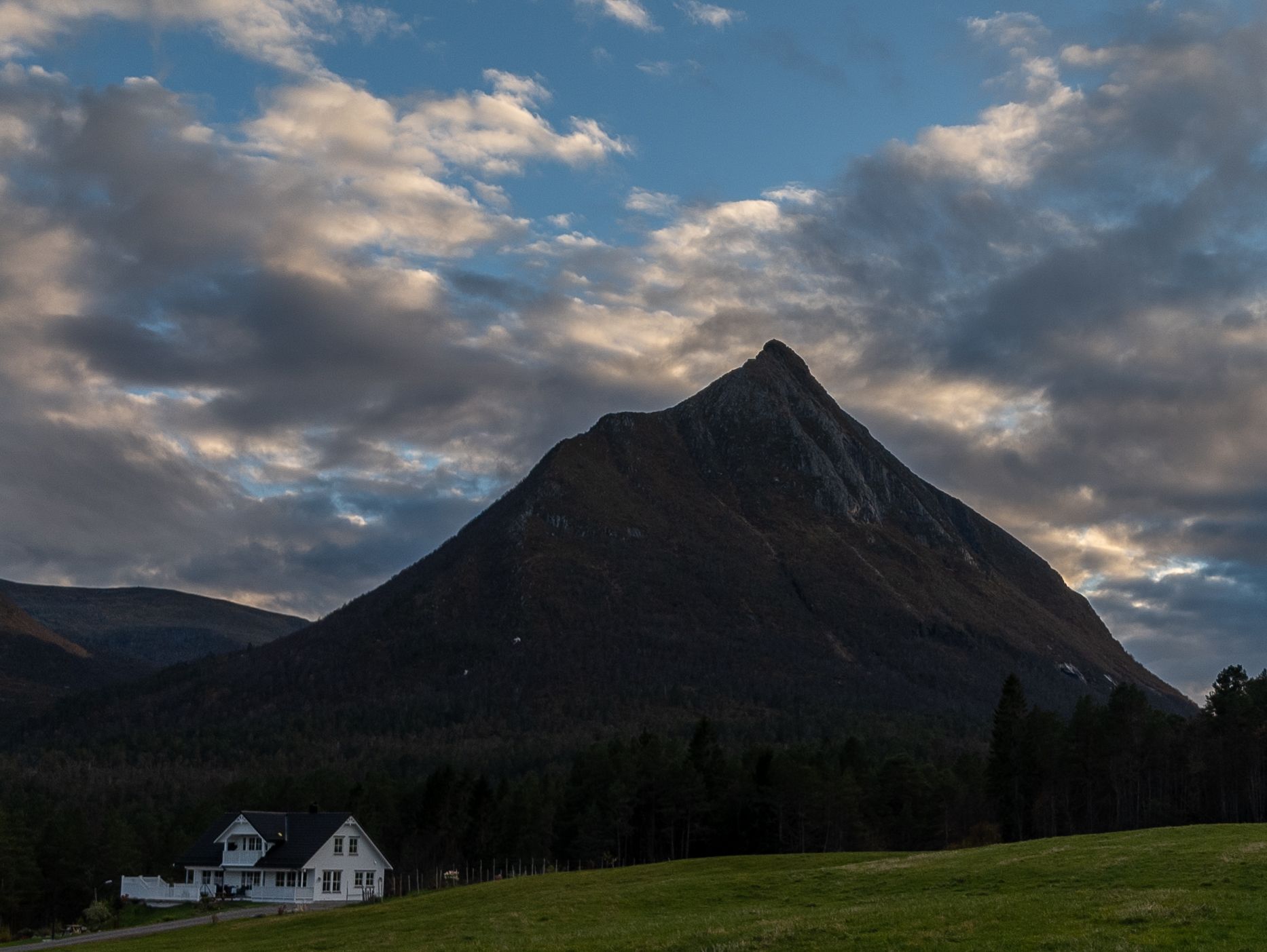 Melens spisse profil over skog, et hvitt hus og grønne jorder under en skyet himmel.