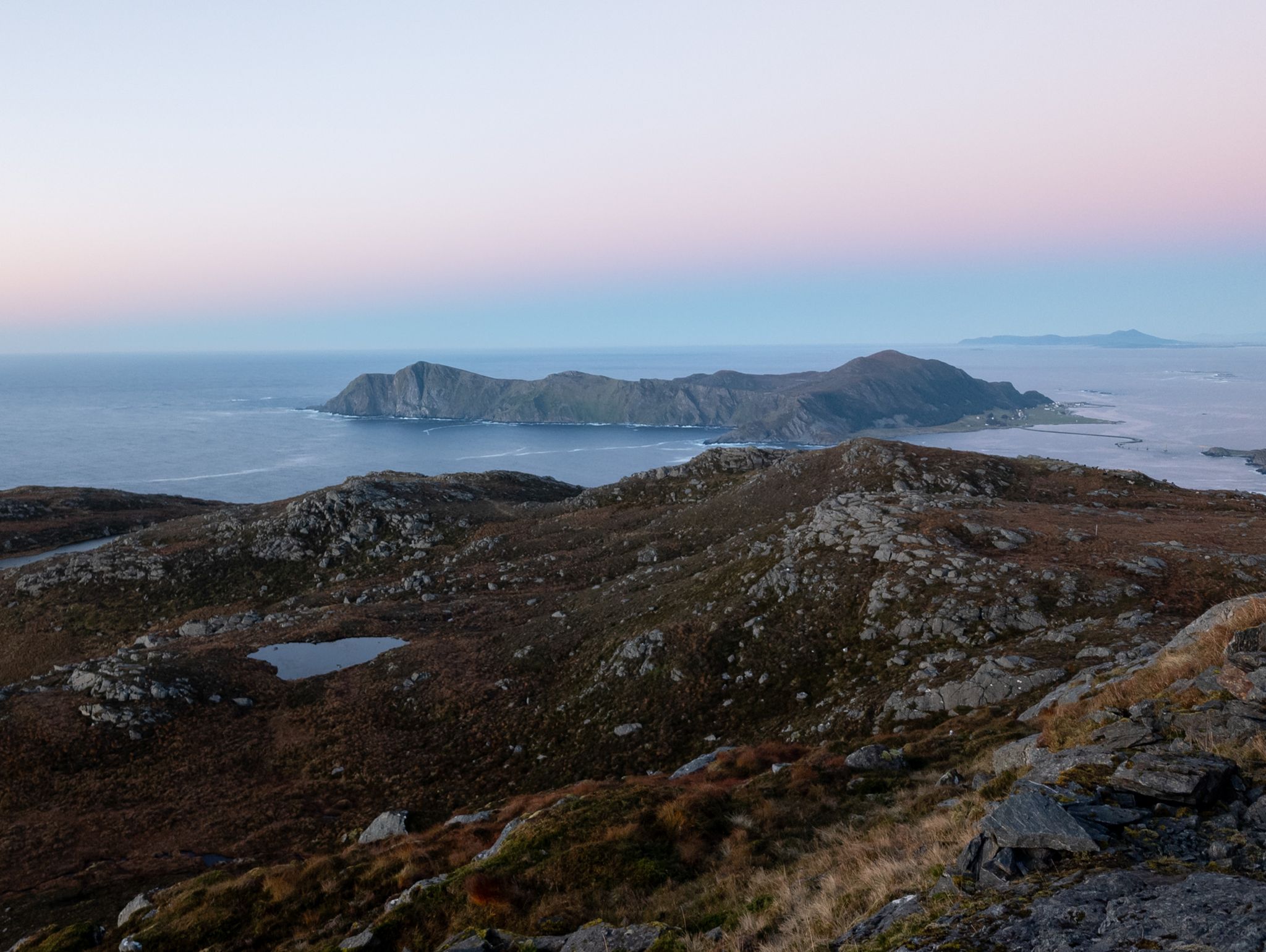 Åpent fjellterreng med små vann, en langstrakt øy og hav under en lys kveldshimmel.