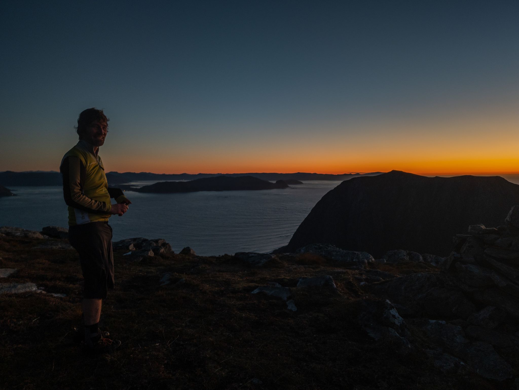 En turkamerat på en steinete topp med øyer og oransje solnedgang over havet bak seg.