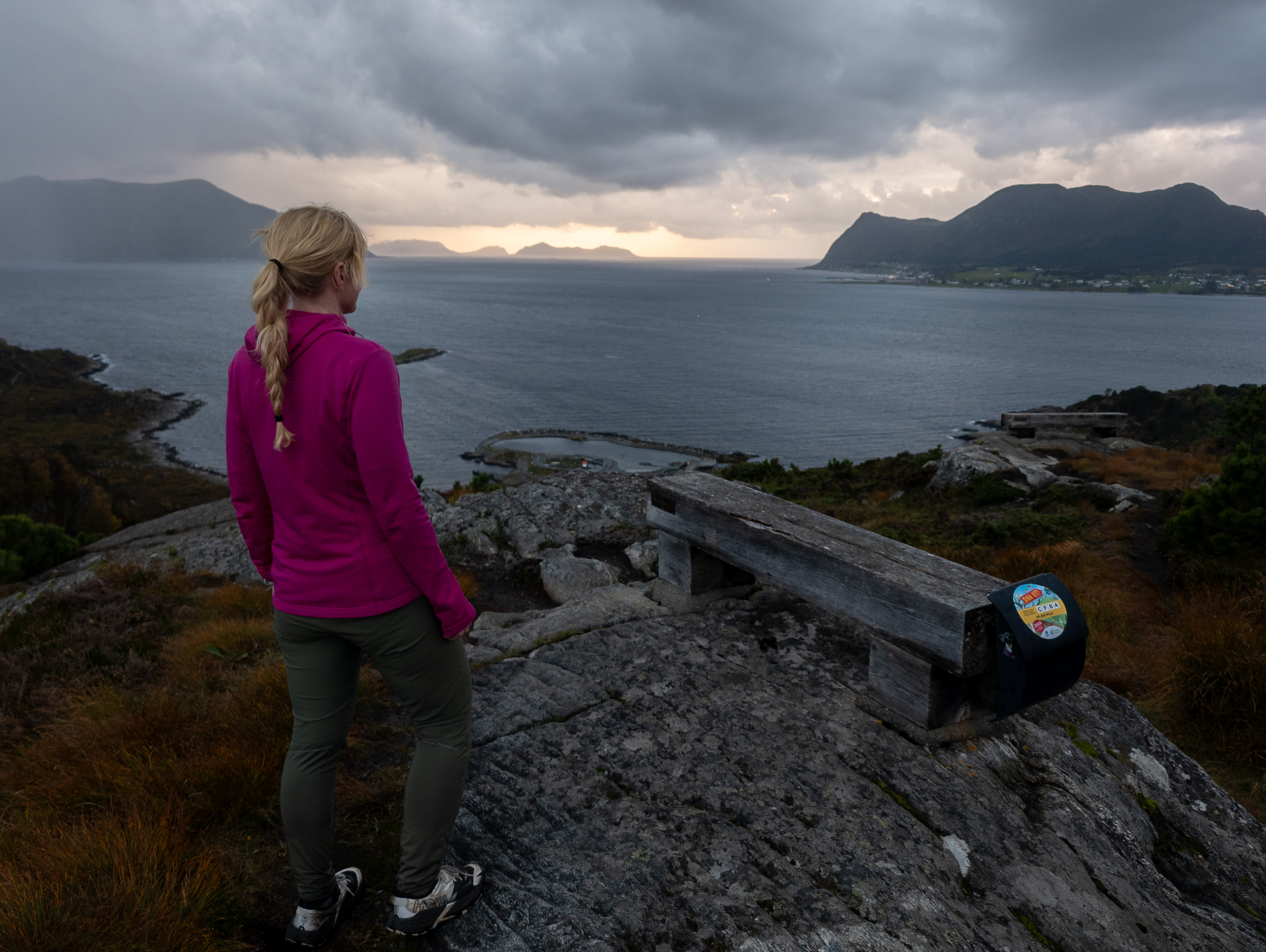 En turgåer i rosa jakke ved en benk på svaberg, med utsikt over sjøen og fjellene i kveldslyset.